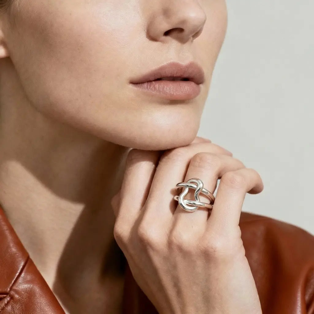 exaequo jewelry A close-up of a model's hand and lower face wearing a silver ring with an abstract, intertwined knot-like design. The model is wearing a brown leather jacket against a neutral light gray background.